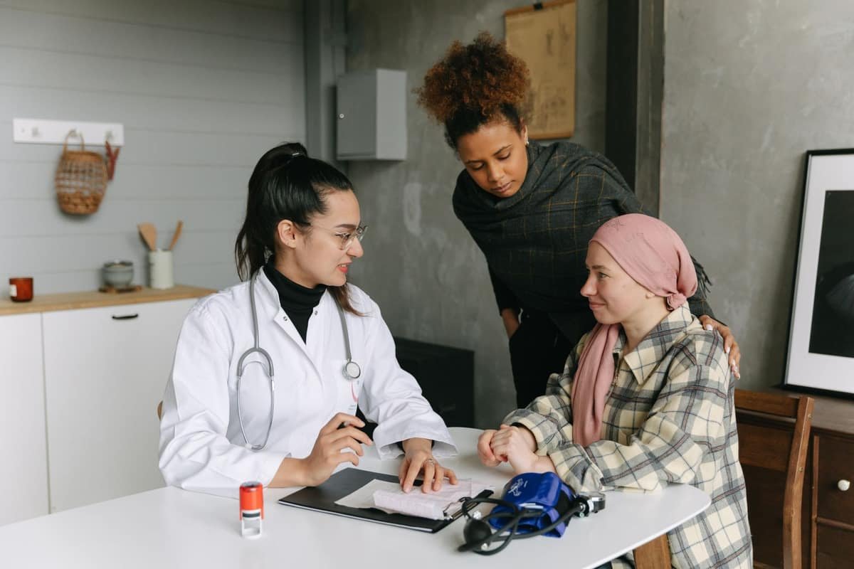 Cancer patient wearing a soft pink head scarf sitting at a home table, hand resting on her shoulder from a supportive companion, in a calm conversation with a doctor
