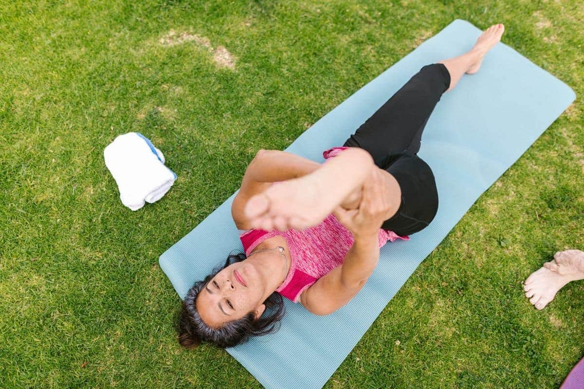 Mature woman lying on a yoga mat outdoors gently pulling one knee toward her chest for a leg stretch